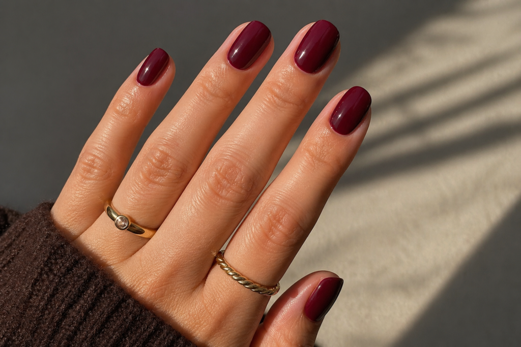 Close-up of glossy dark berry nails in warm sunlight with gold rings and a rich, elegant manicure aesthetic.