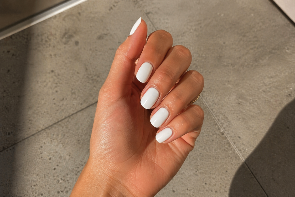 Close-up of glossy milky white nails in natural sunlight with a soft, clean manicure aesthetic and minimal gold jewellery.
