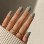 Close-up of glossy sage green nails with gold rings and a cosy cream knit sleeve in soft natural sunlight.